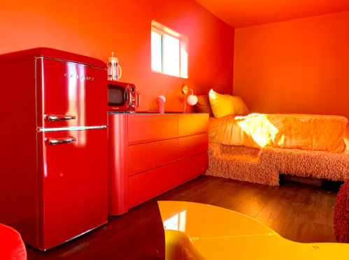 A vibrant orange room featuring a red fridge, a yellow table, and a cozy bed with fluffy bedding.