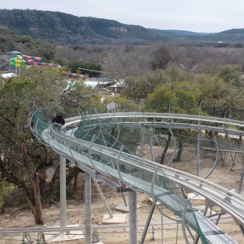A winding roller coaster track surrounded by trees, with a colorful amusement park in the background.