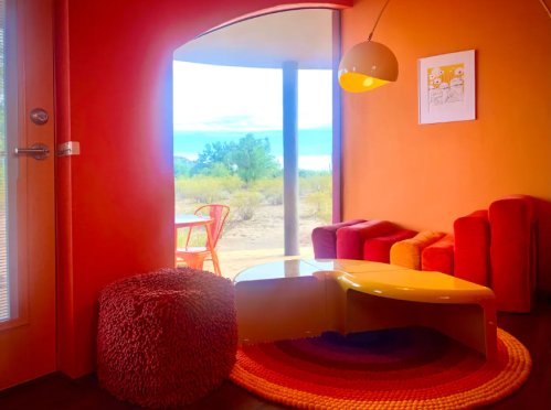 A cozy, brightly colored living space with orange walls, a round table, and a view of the outdoors through a large window.