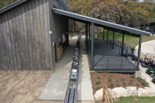 Aerial view of a building with a sloped roof and a track for a ride extending from it, surrounded by grass and rocks.
