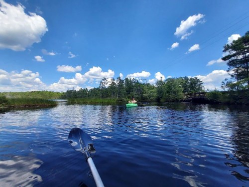 A serene lake scene with a person in a green kayak, surrounded by trees and fluffy clouds in a blue sky.