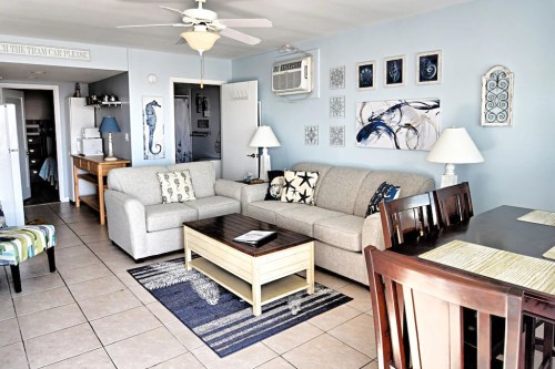 Bright living room with a gray sofa, wooden coffee table, dining area, and beach-themed decor on light blue walls.