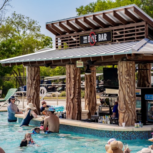 A poolside bar with wooden pillars, surrounded by people enjoying the water on a sunny day.