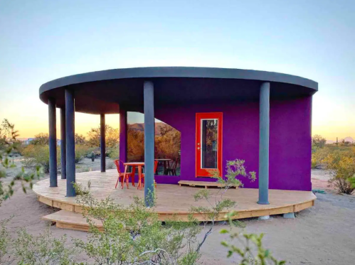 A colorful round house with a purple exterior and orange door, surrounded by desert landscape at sunset.