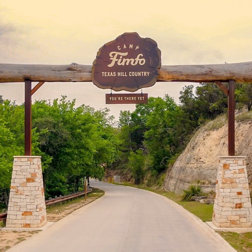 Sign at Camp Fimfo in Texas Hill Country, welcoming visitors with a scenic road lined by trees.