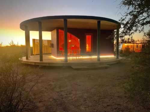 A modern round cabin with glowing lights, surrounded by desert landscape at sunset.
