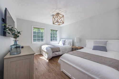 Bright, modern bedroom with two beds, a TV, and natural light from windows. Neutral decor and wooden flooring.