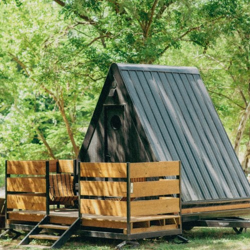 A modern A-frame cabin with a metal roof, surrounded by greenery and featuring a wooden deck.
