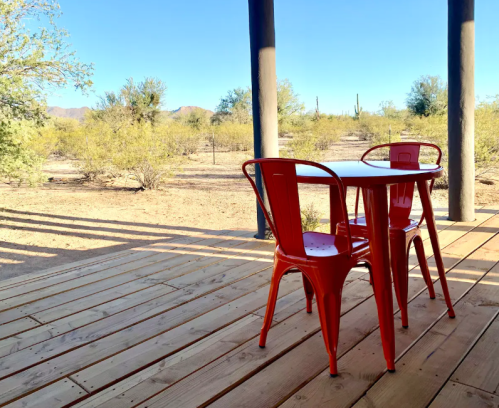 A small red table and chairs on a wooden deck, surrounded by desert vegetation and clear blue skies.