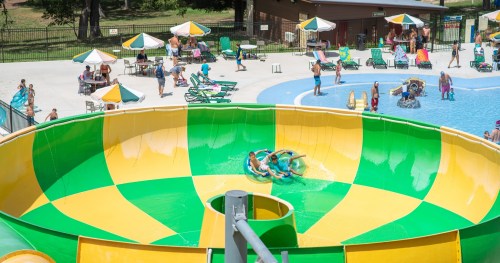 A colorful water slide at a pool, with people enjoying the water and lounging on chairs under umbrellas.