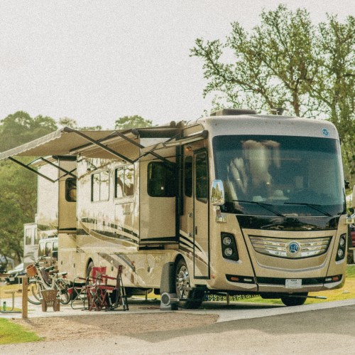 A parked RV with an extended awning, surrounded by trees and outdoor furniture in a campsite setting.