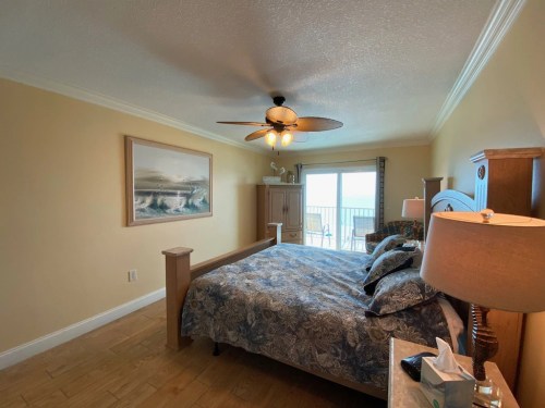 Cozy bedroom with a large bed, ceiling fan, and balcony view, featuring light-colored walls and wooden furniture.