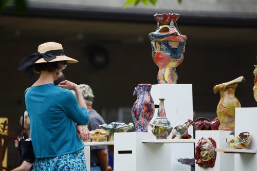 A woman in a straw hat examines colorful pottery displayed on white pedestals at an outdoor art event.