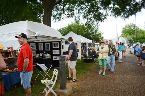 A bustling outdoor market with vendors under tents, people browsing art, and trees lining the pathway.