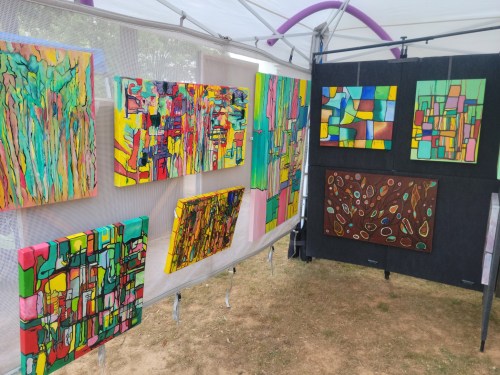 A vibrant art display featuring colorful abstract paintings on canvas, arranged in a tent at an outdoor event.