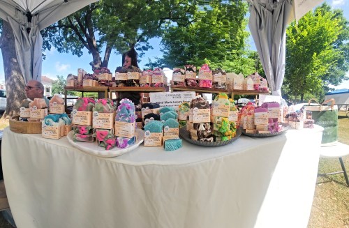 A market stall displaying colorful handmade soaps arranged on wooden trays, with a vendor in the background.