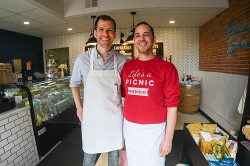 Two smiling men in aprons stand in a cozy bakery, surrounded by food displays and a welcoming atmosphere.