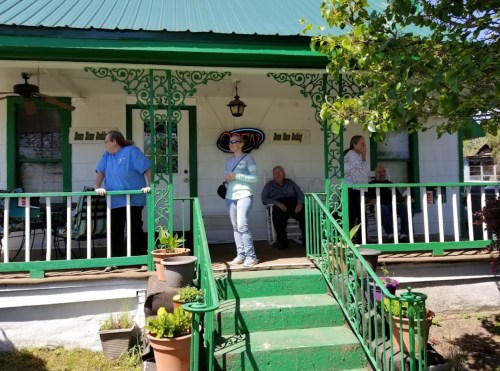 A group of people stands and sits on a porch with green railings, surrounded by potted plants on a sunny day.