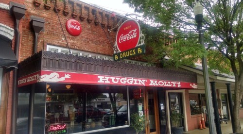 A vintage storefront with a red Coca-Cola sign and "Huggin' Molt's" above the entrance, surrounded by trees.