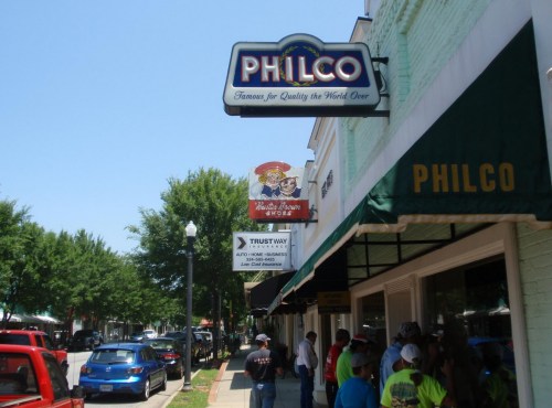 A street view featuring the Philco sign and various storefronts on a sunny day, with people walking nearby.