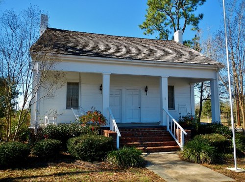 A white, two-story house with a sloped roof, surrounded by greenery and trees under a clear blue sky.