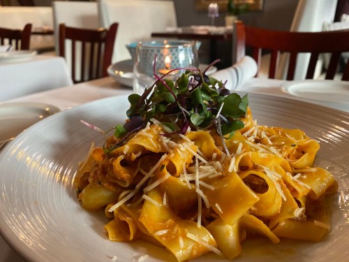 A plate of pasta with ribbons, topped with cheese and microgreens, set on a table with a candle in the background.