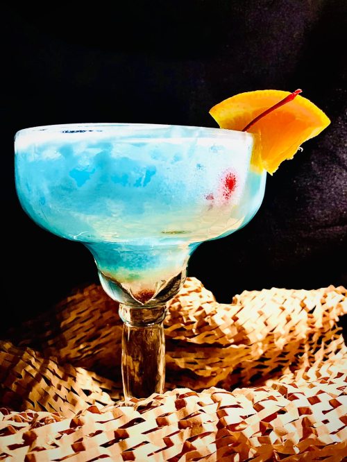 A vibrant blue cocktail in a large glass, garnished with a slice of orange and a cherry, resting on a woven surface.