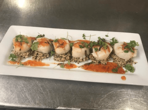 A plate of five scallops on a bed of rice, garnished with herbs and drizzled with a spicy sauce.