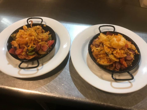 Two plates of paella, one with shrimp and sausage, the other with mixed seafood, served in small black pans.