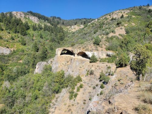 A rocky cliff with a natural arch surrounded by lush greenery and mountains under a clear blue sky.