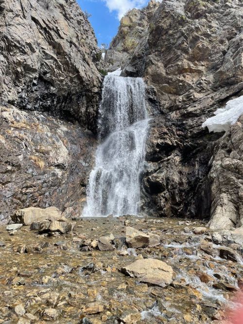 A cascading waterfall flows down rocky cliffs into a clear pool, surrounded by rugged terrain and patches of snow.