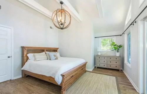 Bright, modern bedroom with a wooden bed, light fixture, dresser, and large windows letting in natural light.