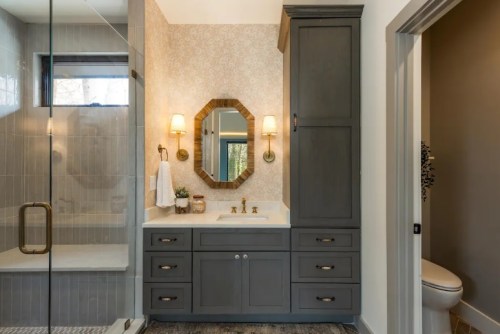 Modern bathroom featuring a gray vanity, gold fixtures, a glass shower, and elegant wall decor.
