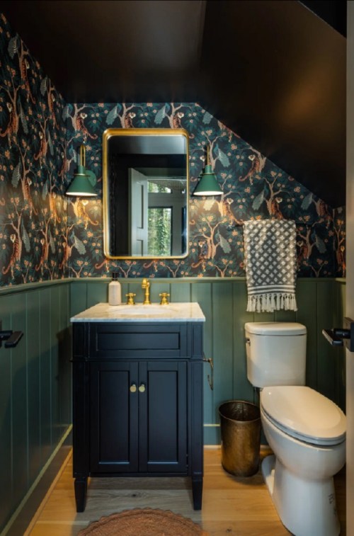 A stylish bathroom featuring a dark vanity, patterned wallpaper, gold fixtures, and a modern toilet under a sloped ceiling.