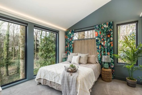 Cozy bedroom with large windows, greenery outside, colorful curtains, and a stylish bed with decorative pillows.
