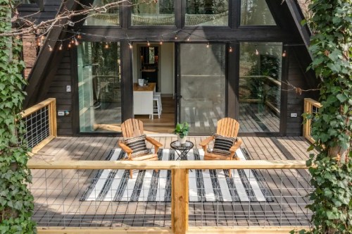 A cozy deck with two wooden chairs, a small table, and string lights, surrounded by greenery and a modern A-frame cabin.