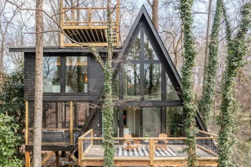 A modern A-frame cabin nestled in trees, featuring large windows and a spacious deck with wooden chairs.
