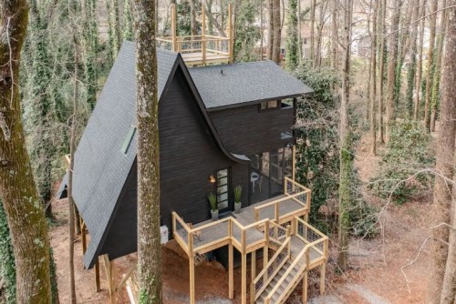 A modern black house nestled in a wooded area, featuring a large deck and surrounded by tall trees.