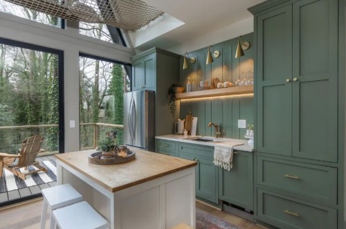 Modern kitchen with green cabinetry, wooden island, and large windows overlooking a forested area.