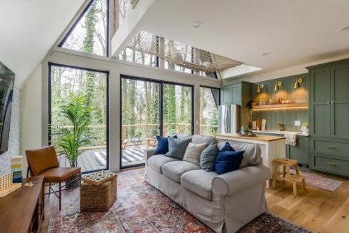 Cozy living room with large windows, green cabinetry, and a view of trees outside, featuring a sofa and wooden accents.