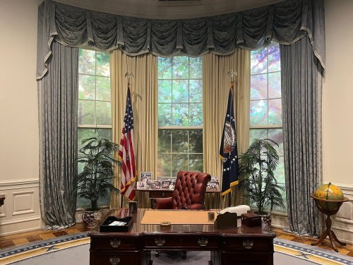 A well-decorated office with a large desk, an armchair, flags, and windows overlooking greenery.