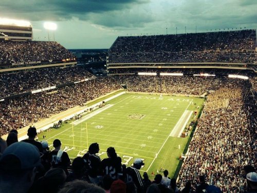 Aerial view of a packed stadium during a football game, with fans cheering and a cloudy sky above.
