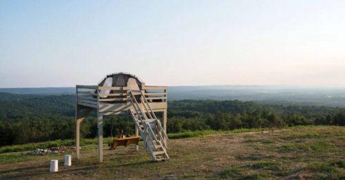 A wooden platform with a tent on top overlooks a vast green landscape under a clear sky.