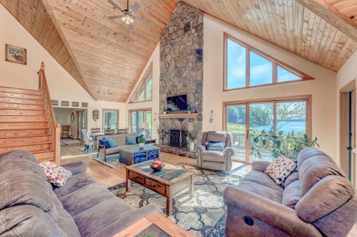 Spacious living room with high ceilings, stone fireplace, large windows, and cozy furniture overlooking a lake.