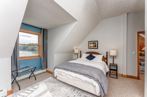 Cozy bedroom with sloped ceiling, window view of water, and simple decor featuring a bed, lamps, and a small table.