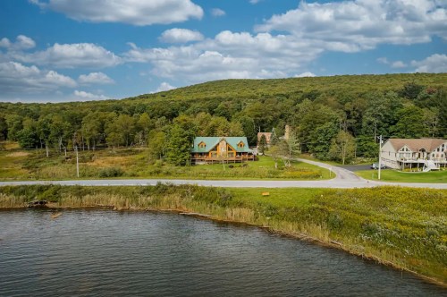 A scenic view of a house by a lake, surrounded by trees and hills under a partly cloudy sky.