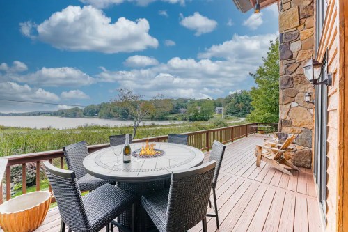 A scenic deck with a round table, chairs, and a fire pit overlooking a calm lake and lush greenery under a blue sky.