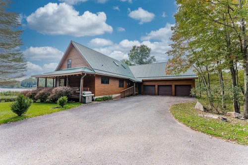A spacious wooden house with a sloped roof, surrounded by greenery and a gravel driveway, under a blue sky with clouds.