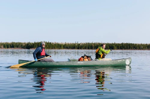 Two people in a canoe on calm water, surrounded by trees and a clear sky, enjoying a peaceful outdoor adventure.
