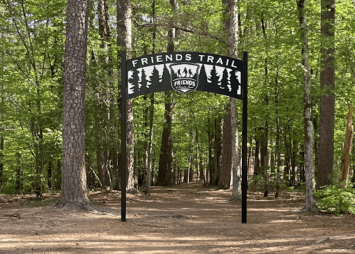 A sign reading "Friends Trail" stands at the entrance of a wooded path surrounded by tall trees.